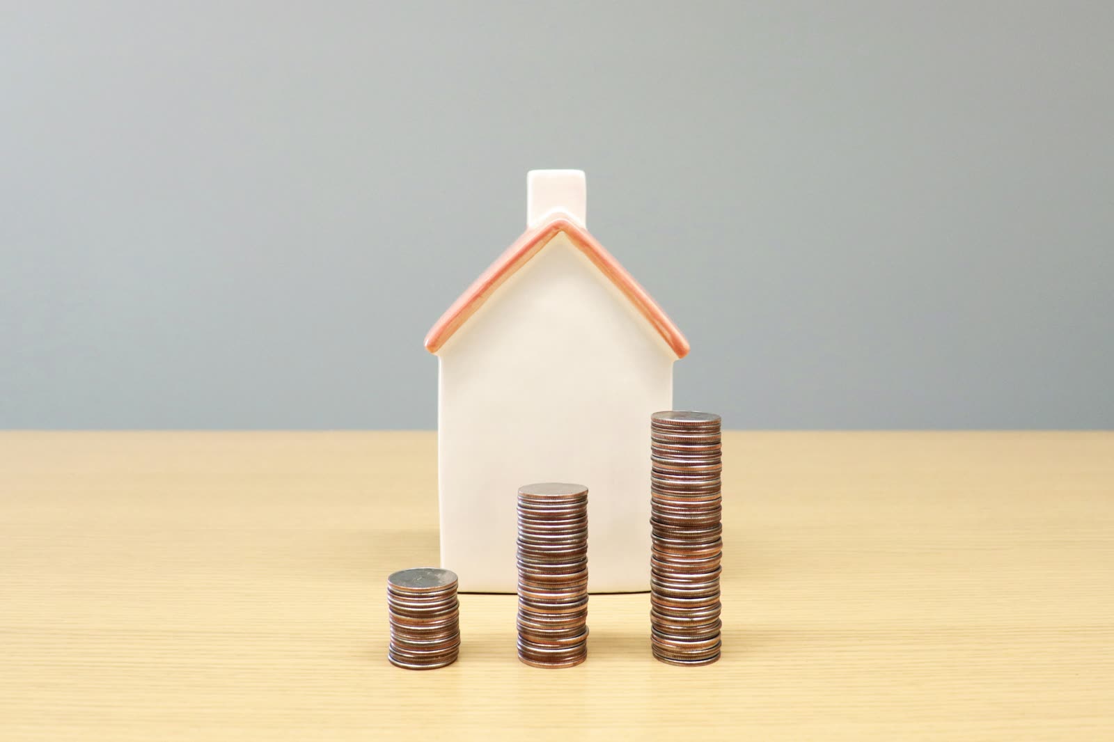 A small wooden model house next to stacks of coins on a desk, representing property taxes and home costs for Michigan buyers.