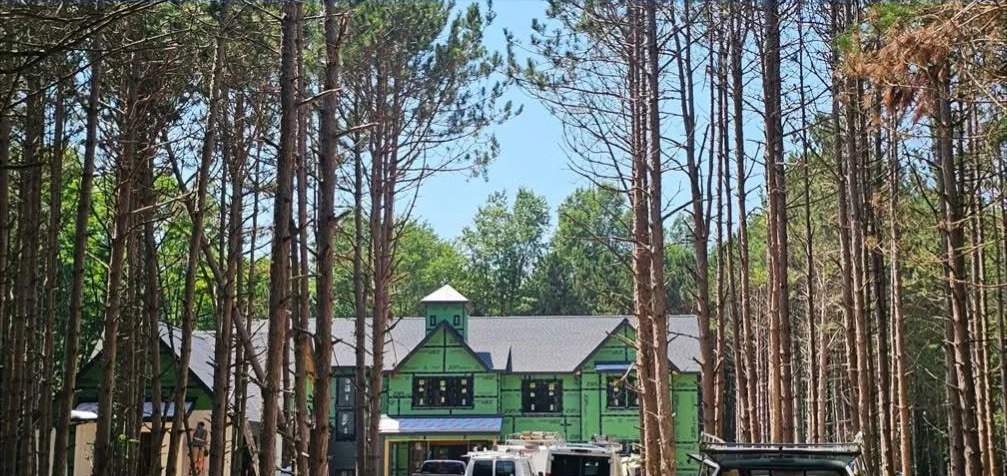A two-story home under construction in green sheathing, set back in a stand of tall pines with work trucks in the drive